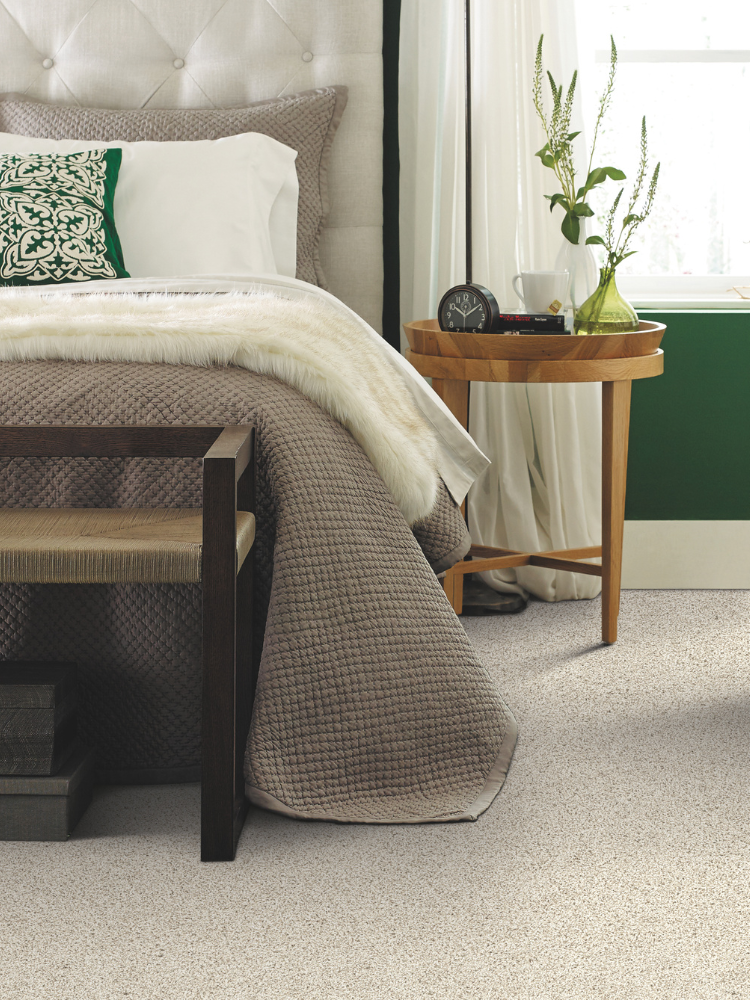 Off-white carpet in bedroom, side table with clock and plants, bed, pillows.
